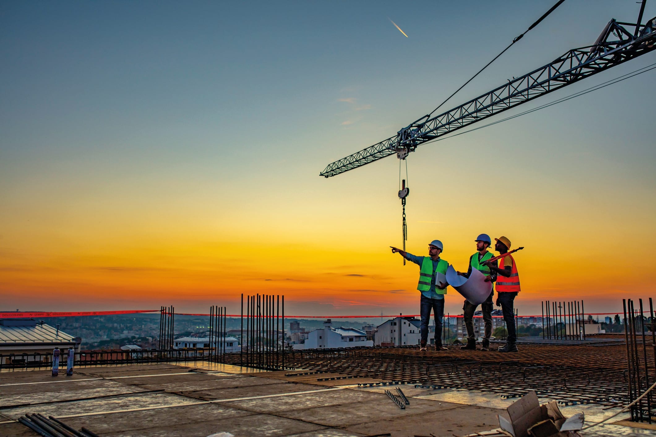Three people in hard hats and high vis vests talking on a rooftop, with an orange sky and crane in the background